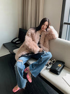 Woman in a fluffy pink coat sitting on a couch in a room with a desk and window.