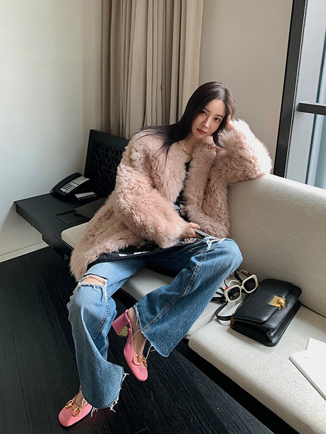 Woman in a fluffy pink coat sitting on a couch in a room with a desk and window.