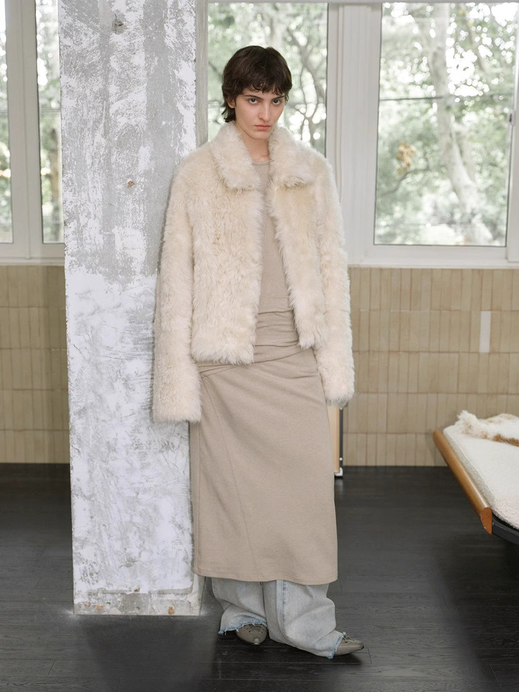 Person wearing a cream fur coat and beige dress in a room with large windows.