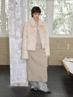 Person wearing a cream fur coat and beige dress in a room with large windows.
