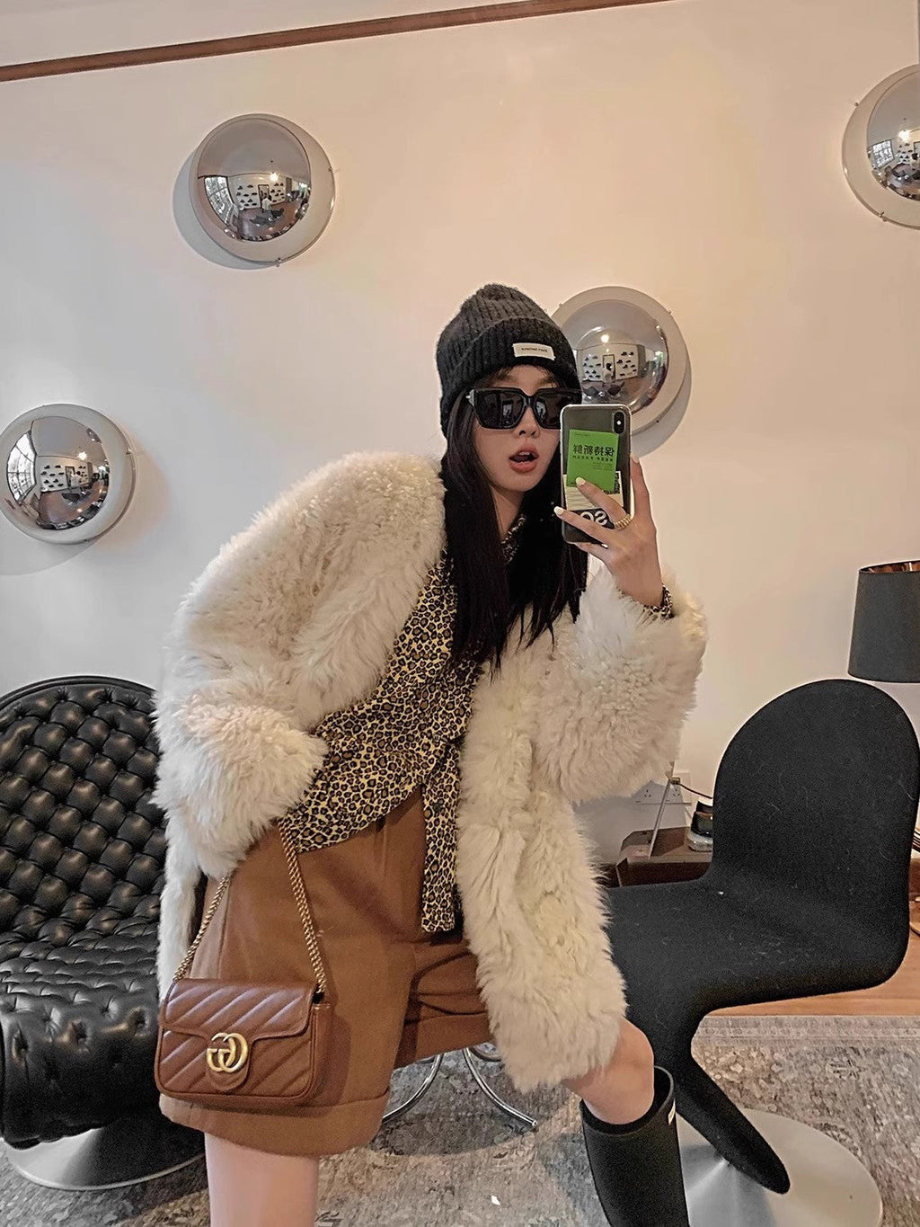 Person taking a mirror selfie wearing a white fur coat, brown bag, and black boots in an indoor setting.