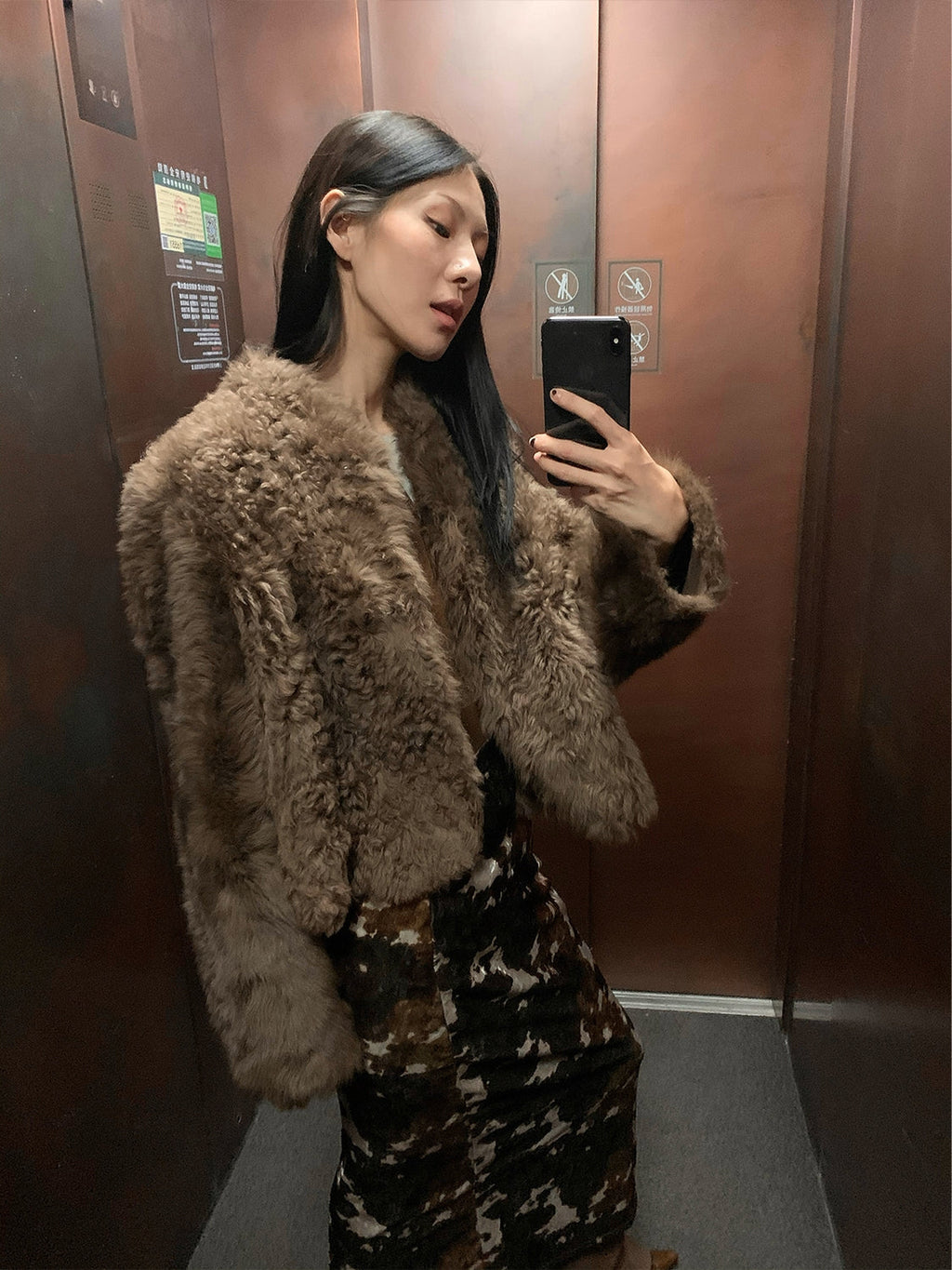 Person wearing a fluffy brown coat taking a mirror selfie in an elevator.