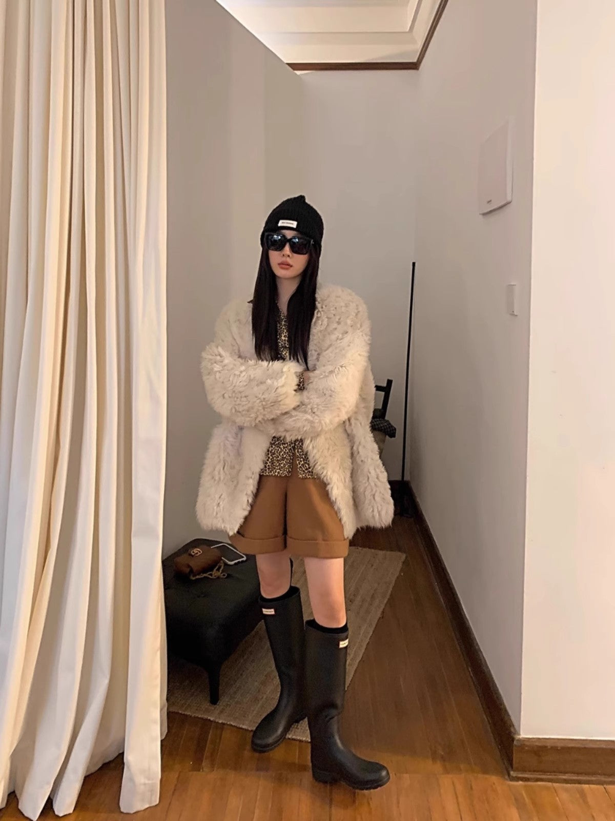 Person wearing a white fluffy coat, brown shorts, black boots, and a black beanie in a room.