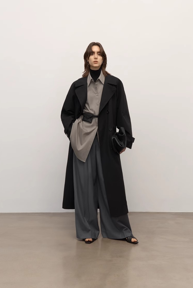 Person wearing a long black coat over a gray outfit on a plain background