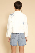 Person wearing a white shirt with blue bow details and denim shorts on a beige background