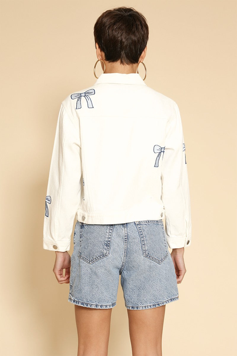 Person wearing a white shirt with blue bow details and denim shorts on a beige background