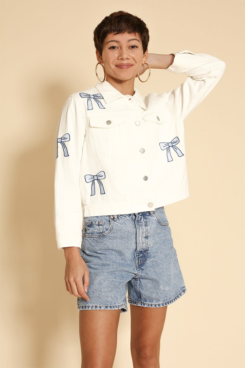 Woman wearing a white denim jacket with blue bow patterns and denim shorts on a beige background