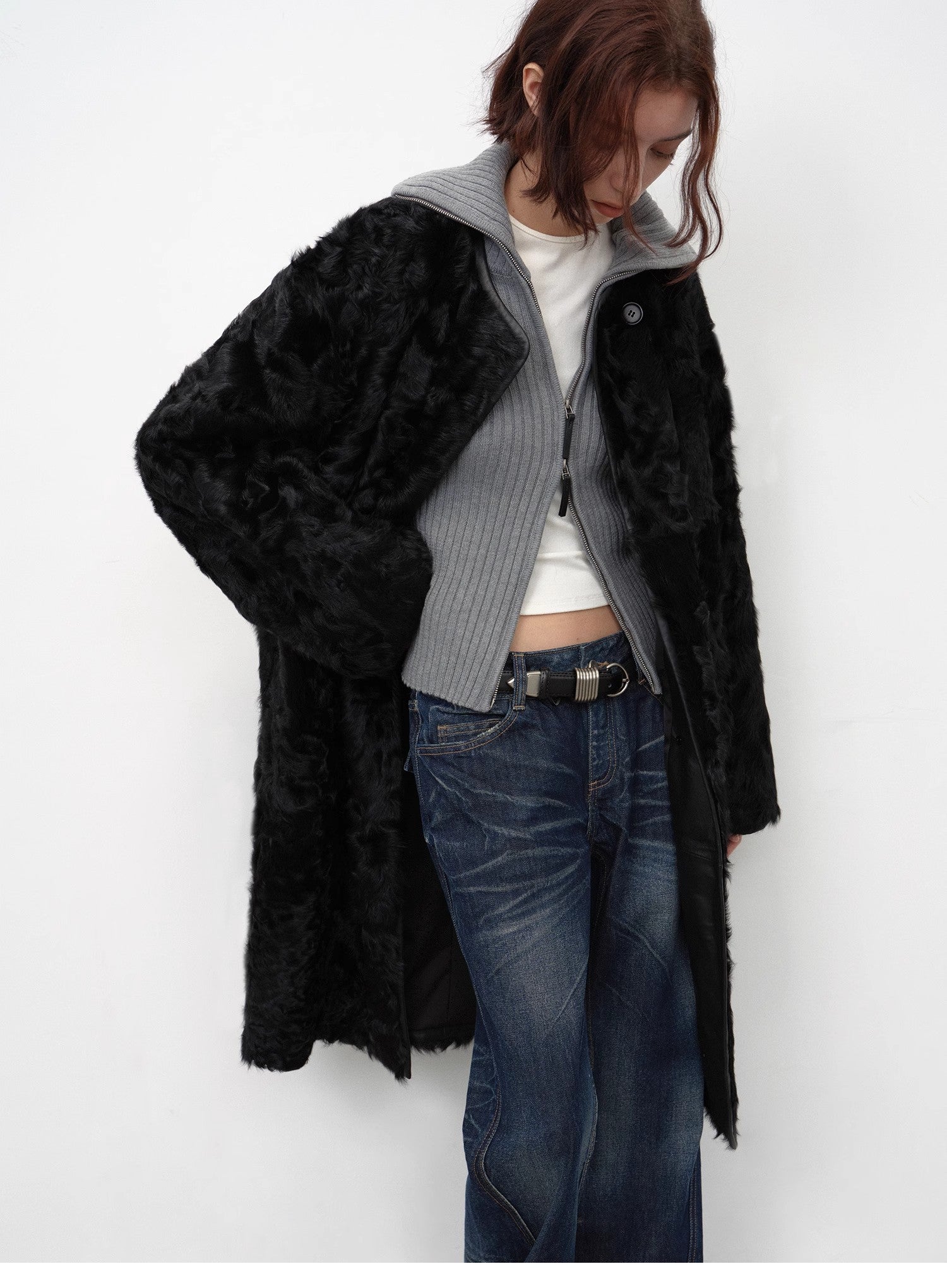Person wearing a black fur coat over a gray sweater with a white shirt, blue jeans, and a belt.