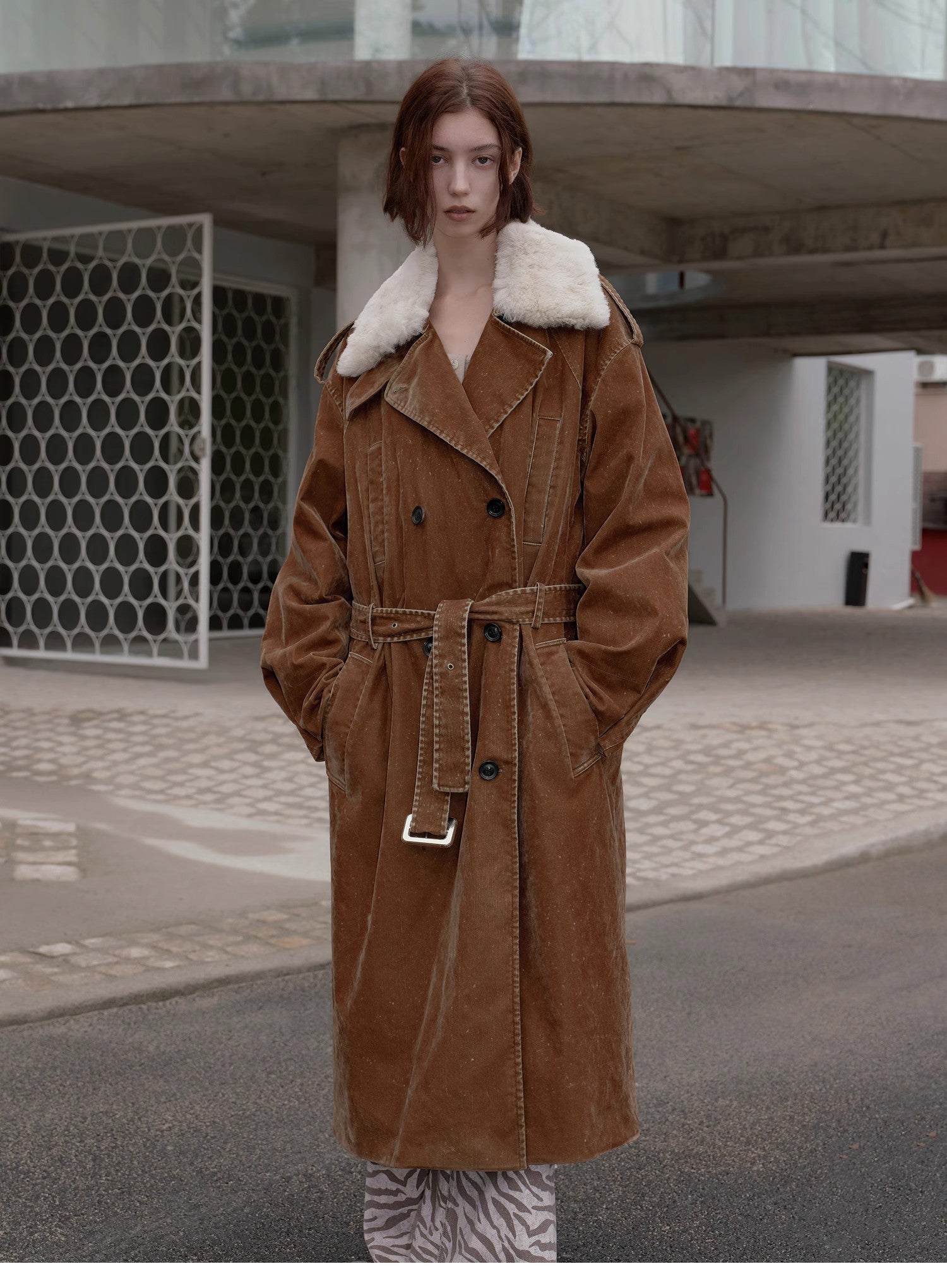 Person wearing a brown coat with white fur collar standing on a street.