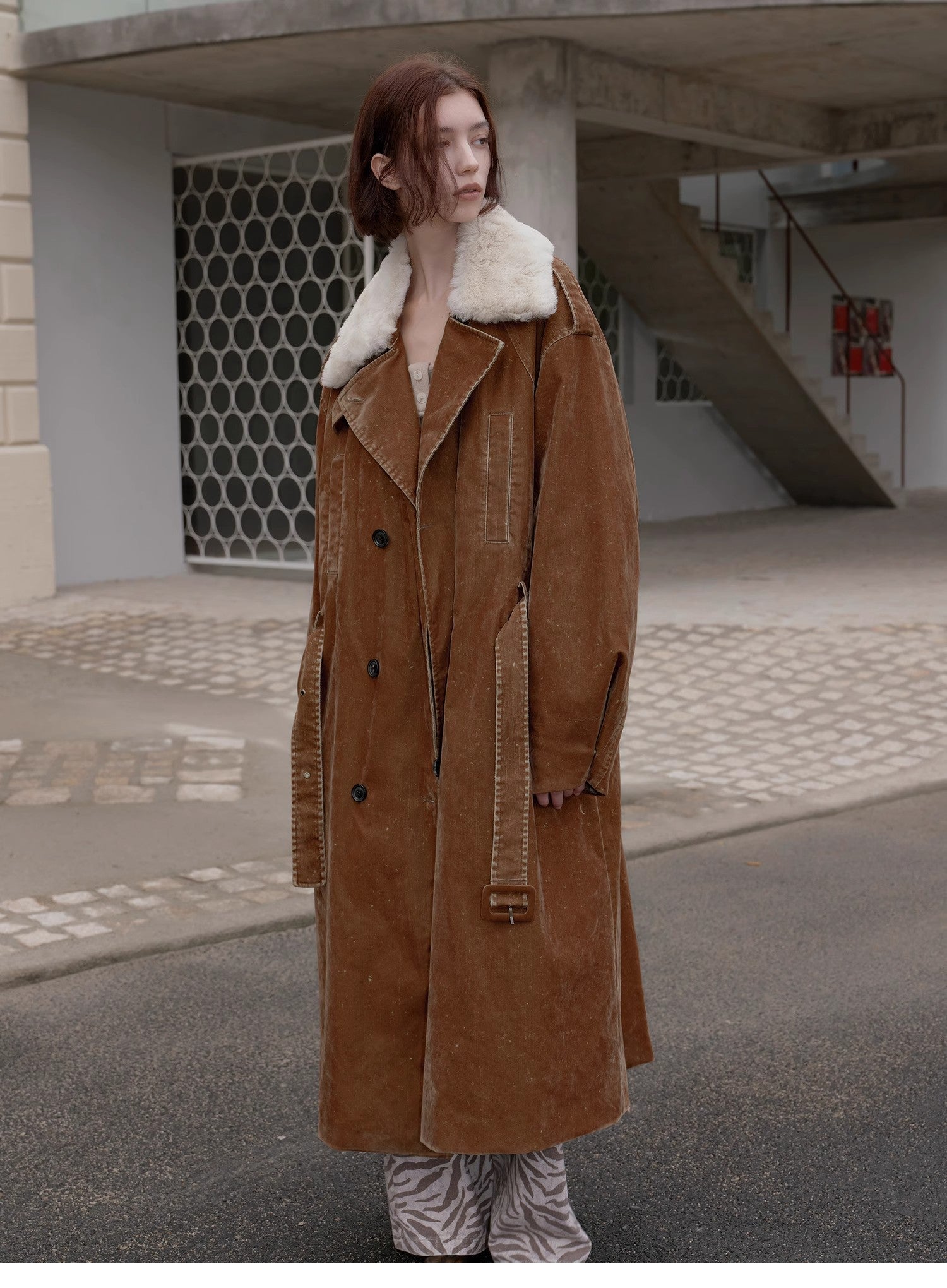 Person wearing a long brown coat with white fur lining in an urban setting