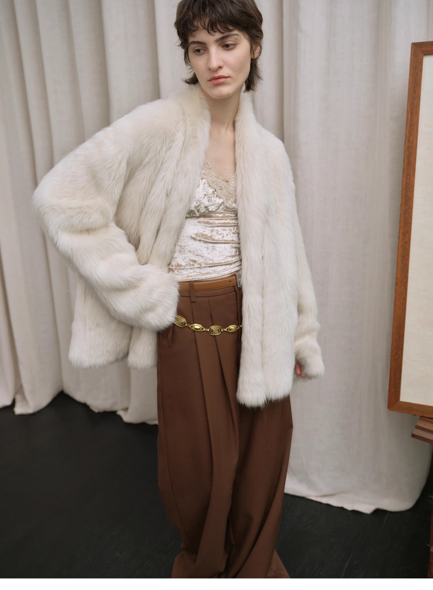 Person wearing a white fluffy coat over a patterned top and brown pants against a neutral background.