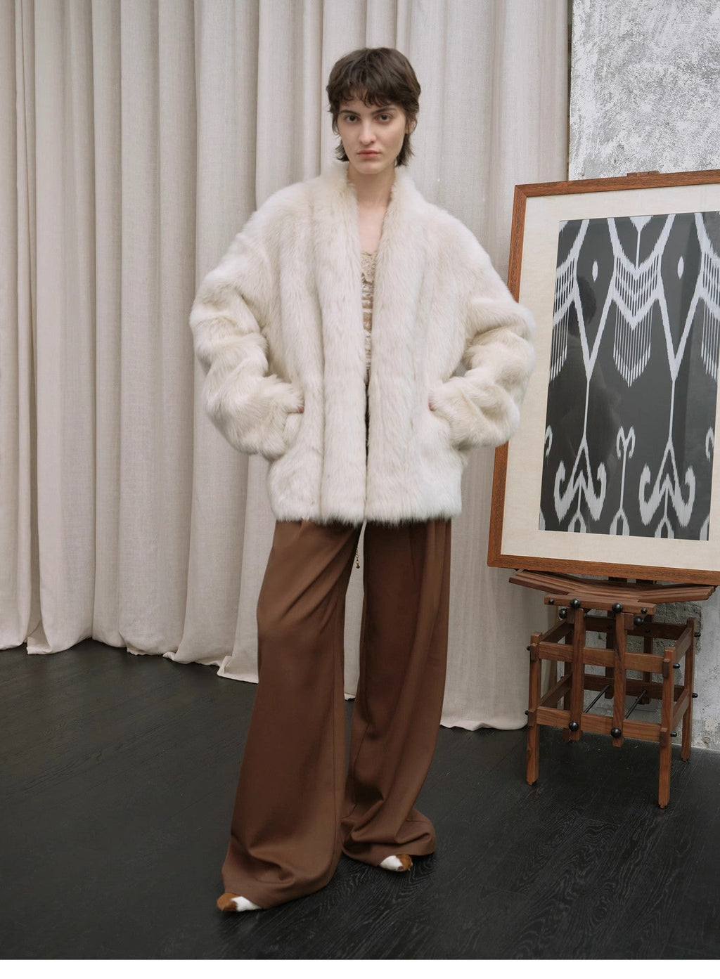 Person wearing a white fur coat and brown pants standing in front of a curtain and an easel with a painting.