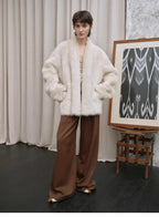 Person wearing a white fur coat and brown pants standing in front of a curtain and an easel with a painting.