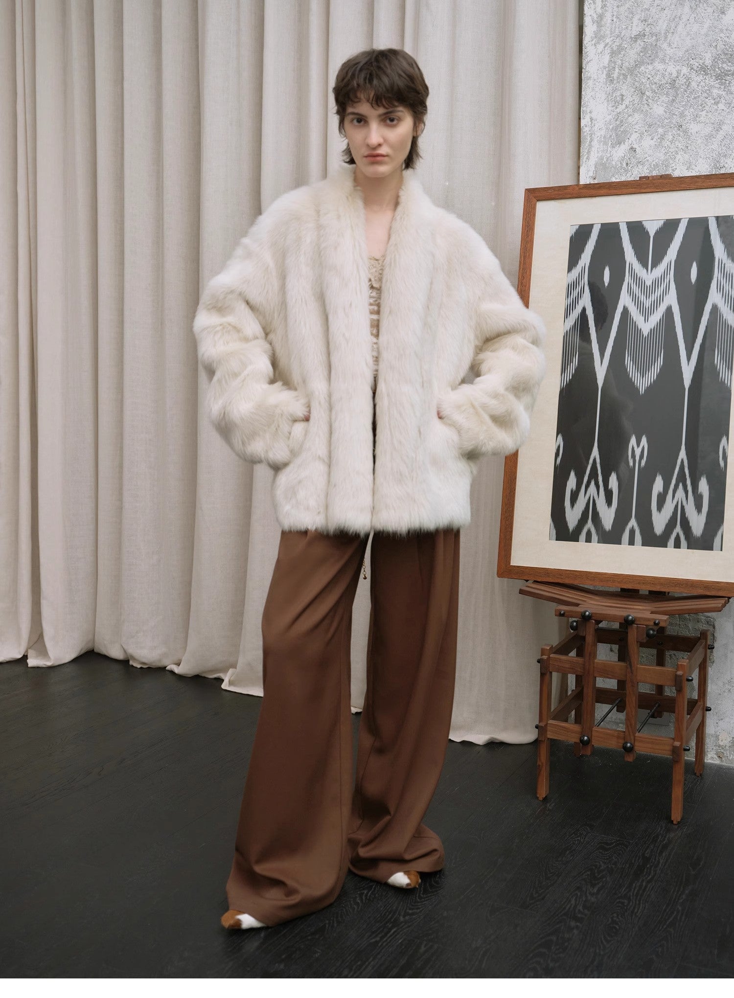 Person wearing a white fur coat and brown pants standing in front of a curtain and an easel with a painting.