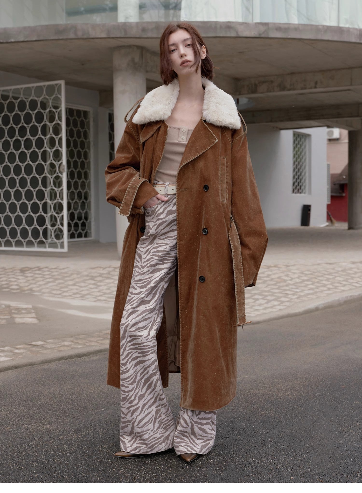 Person wearing a brown coat with white fur lining, beige top, and zebra print pants standing on a street.