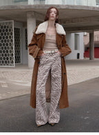Person wearing a brown coat with white fur trim, beige top, and zebra print pants standing on a street.