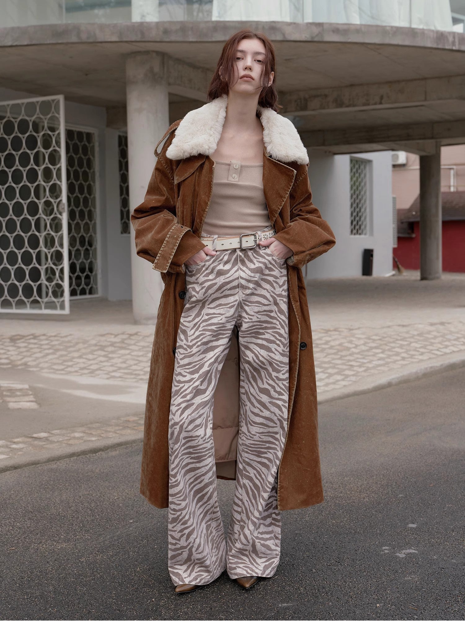 Person wearing a brown coat with white fur trim, beige top, and zebra print pants standing on a street.