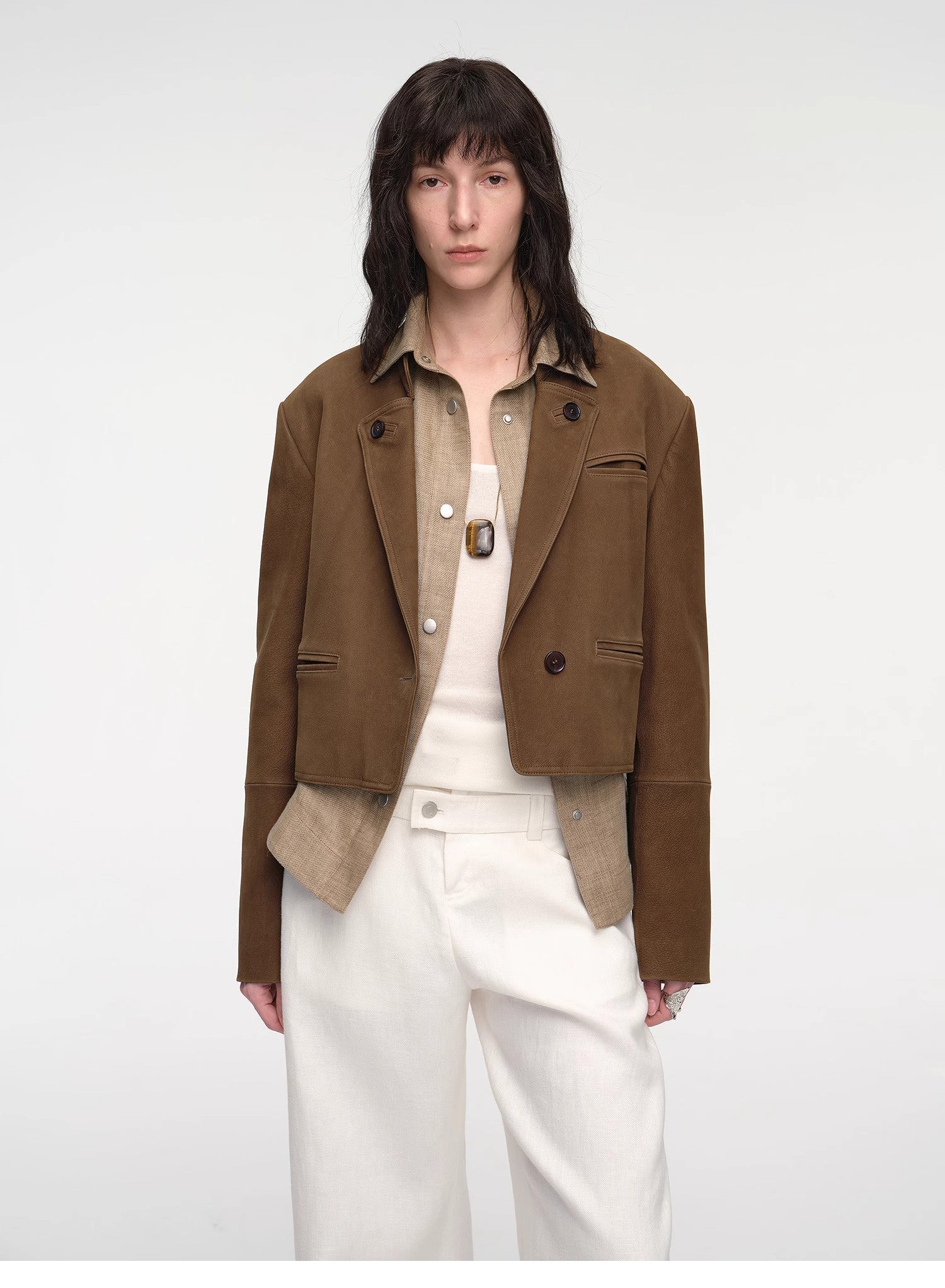 Person wearing a brown jacket over a beige shirt with white pants on a white background