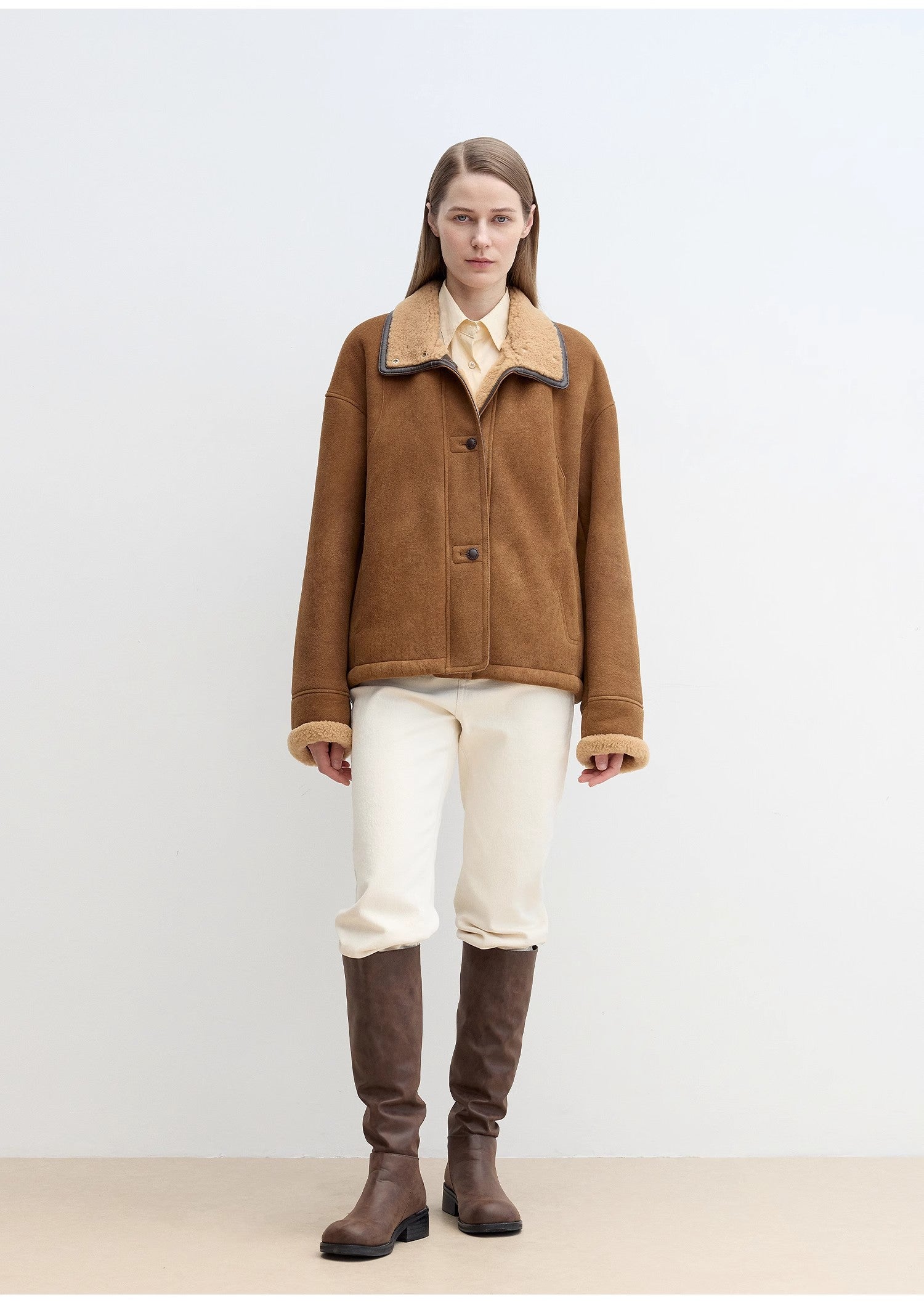 Person wearing a brown jacket with a fur collar and white pants on a light background