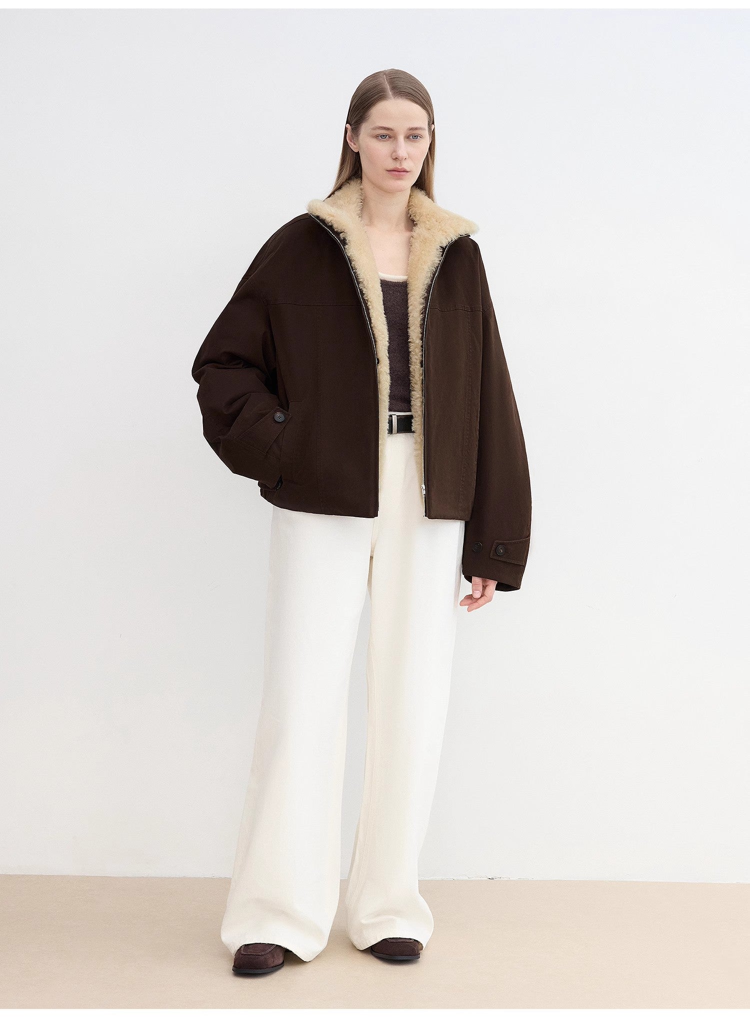 Person wearing a brown jacket with shearling collar and white pants on a plain background