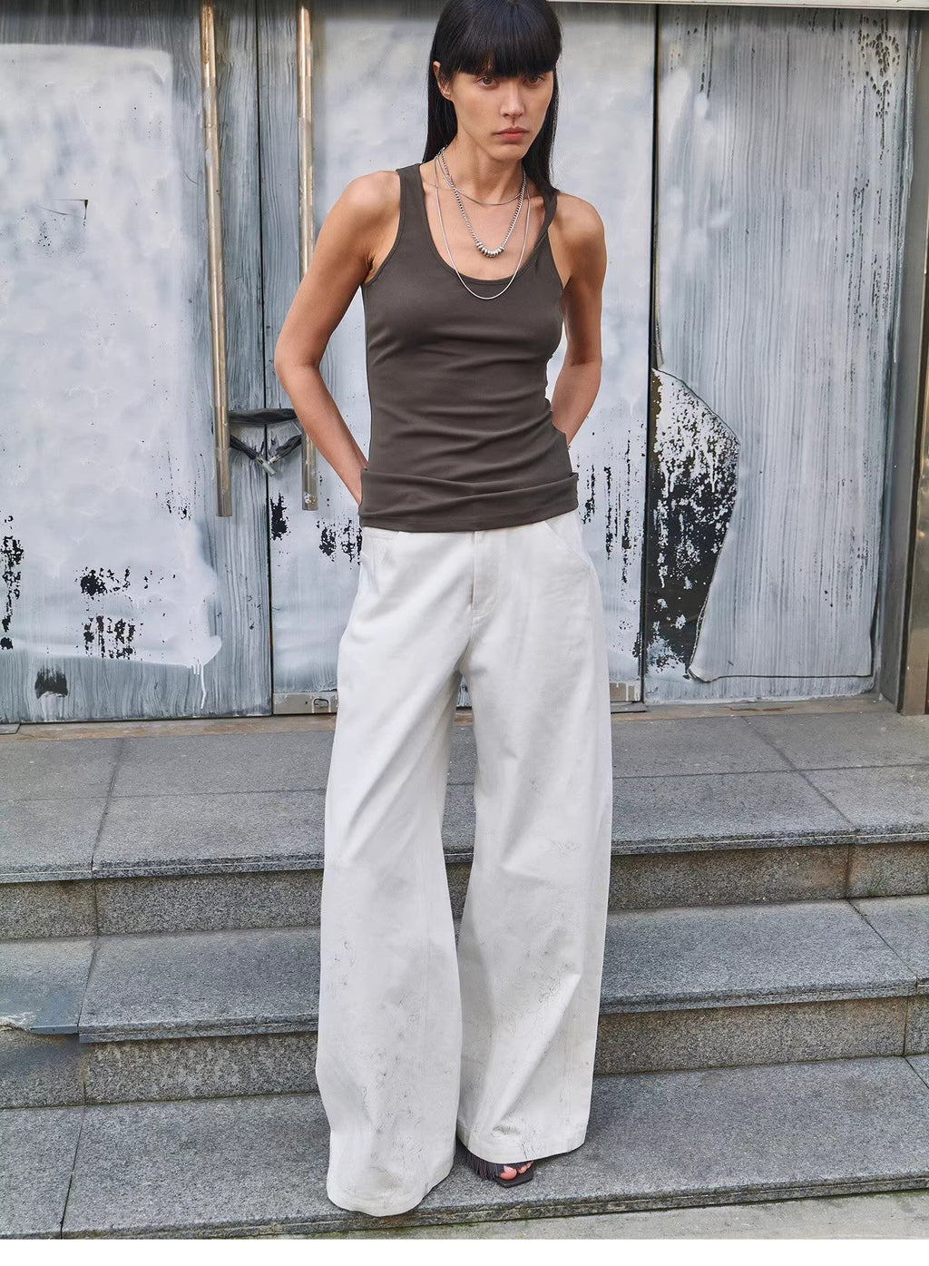 Woman wearing a brown tank top and white pants standing on steps with a textured wall background