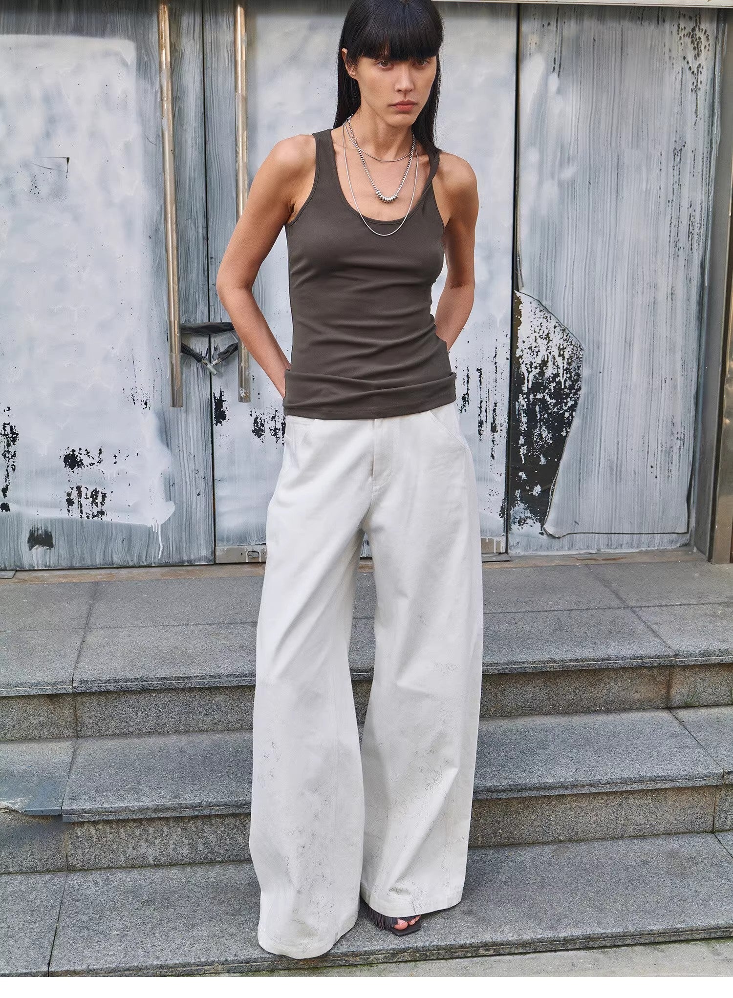 Woman wearing a brown tank top and white pants standing on steps with a textured wall background