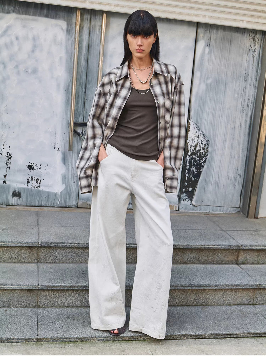 Woman wearing a plaid shirt, brown top, and white pants standing on steps with a textured wall background.