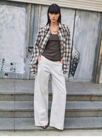 Woman wearing a plaid shirt, brown top, and white pants standing on steps with a textured wall background.