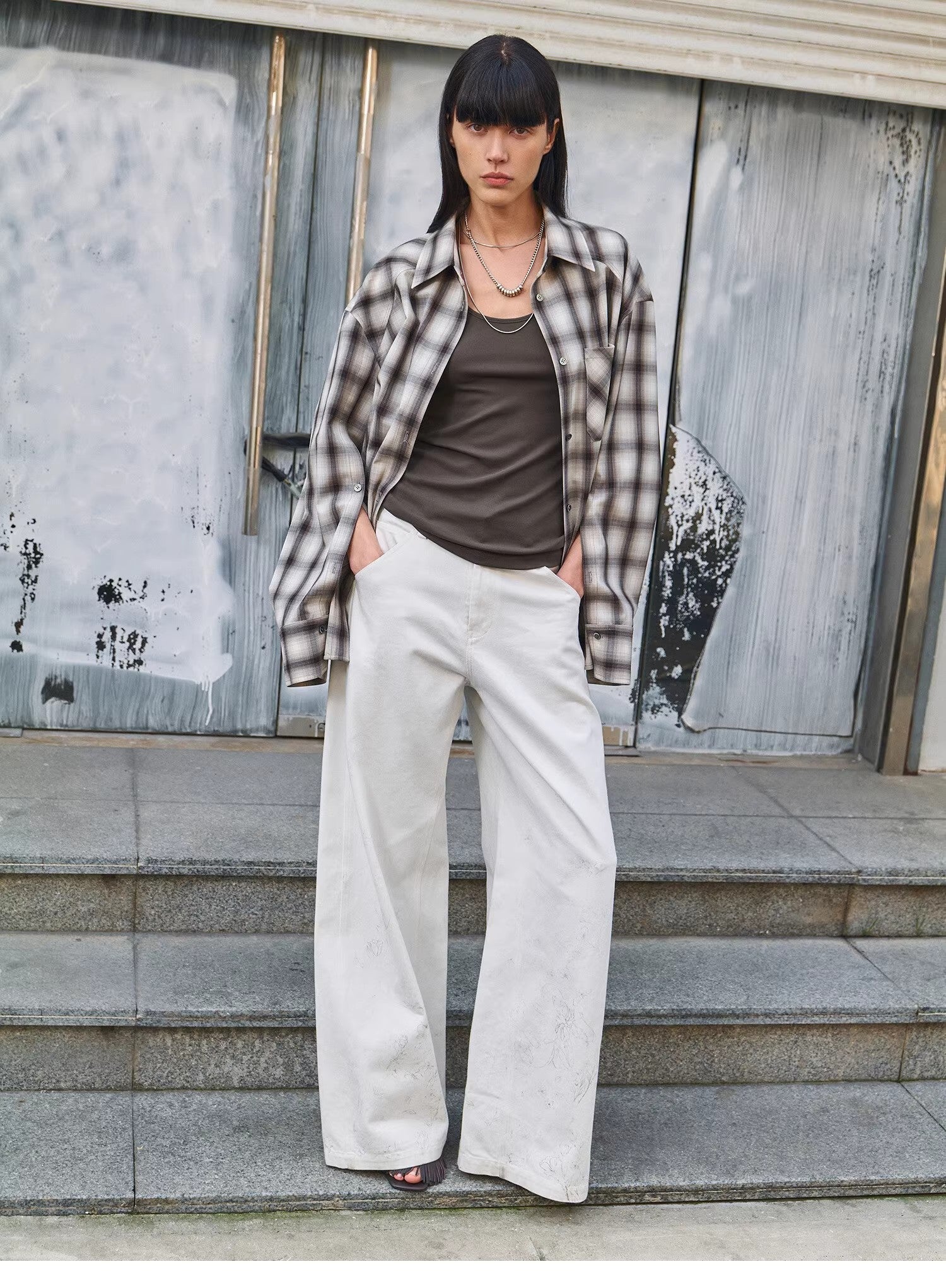 Woman wearing a plaid shirt, brown top, and white pants standing on steps with a textured wall background.