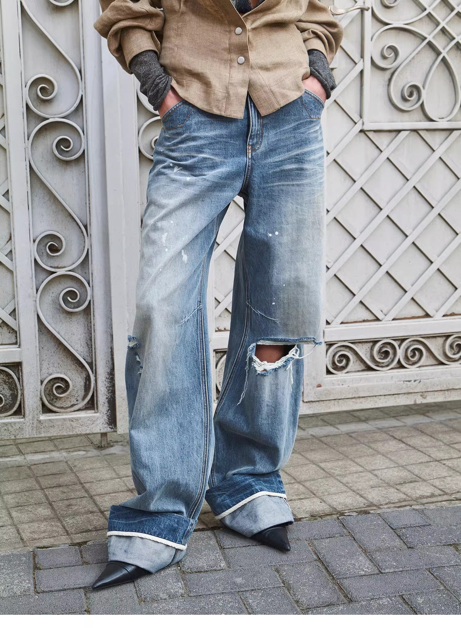 Person wearing a beige shirt and blue jeans standing against an ornate metal gate.