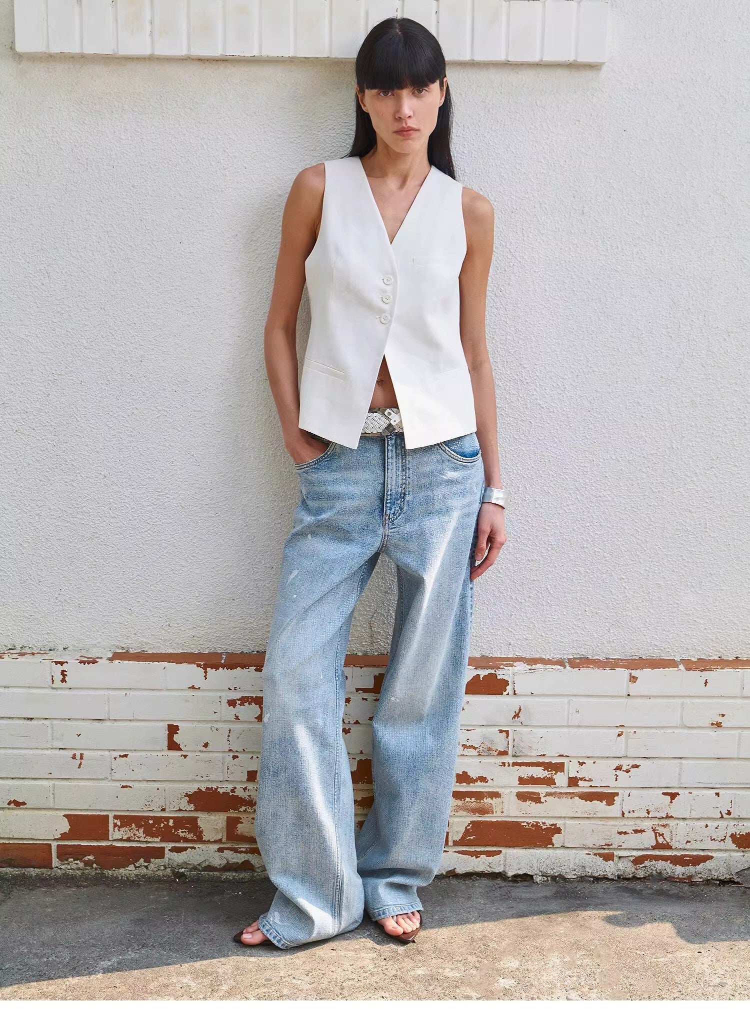 Woman wearing a white sleeveless top and blue jeans standing against a textured wall.