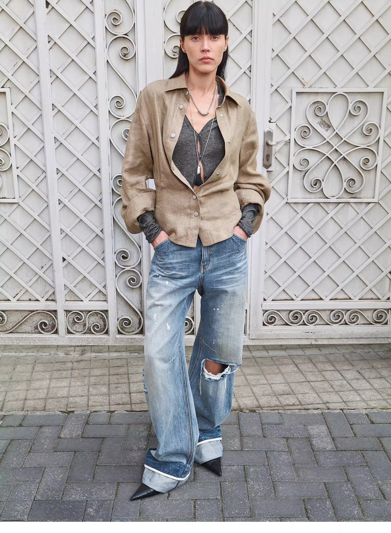 Person wearing a beige jacket, black top, and ripped jeans standing in front of an ornate metal gate.