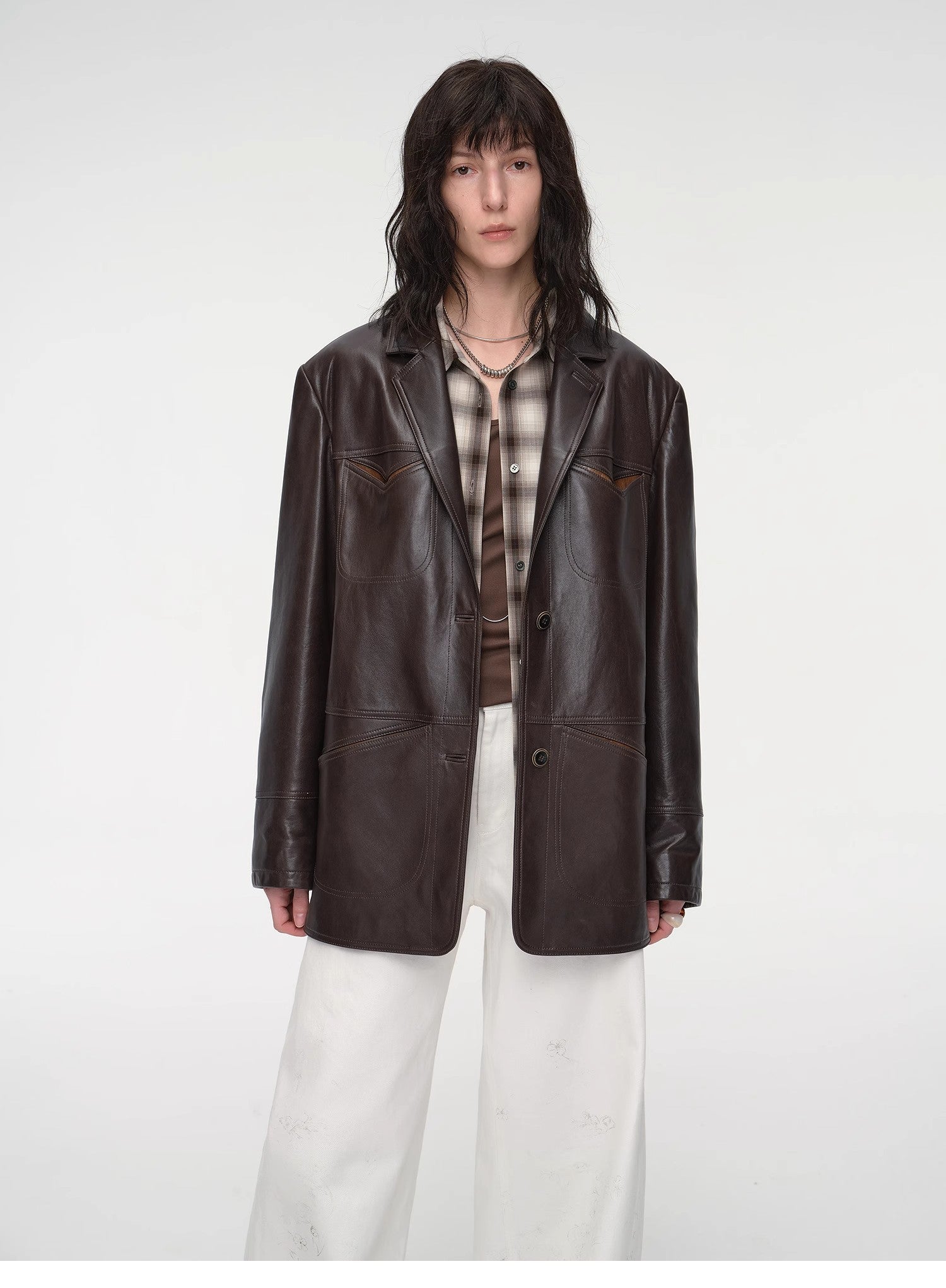 Person wearing a brown leather jacket over a plaid shirt with white pants on a white background