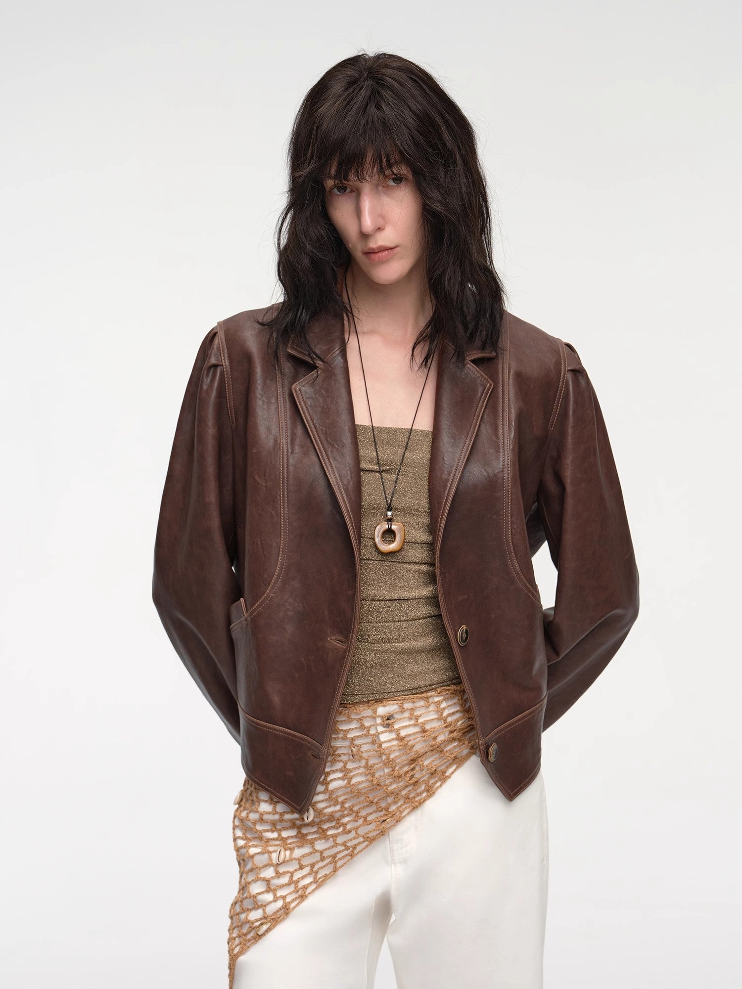 Woman wearing a brown leather jacket over a patterned top and white pants on a plain background