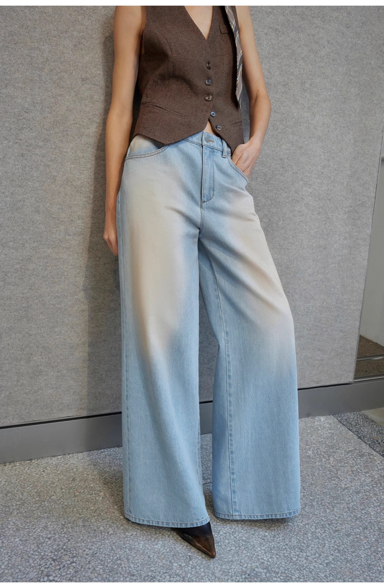 Person wearing a brown vest and light blue jeans against a gray wall.