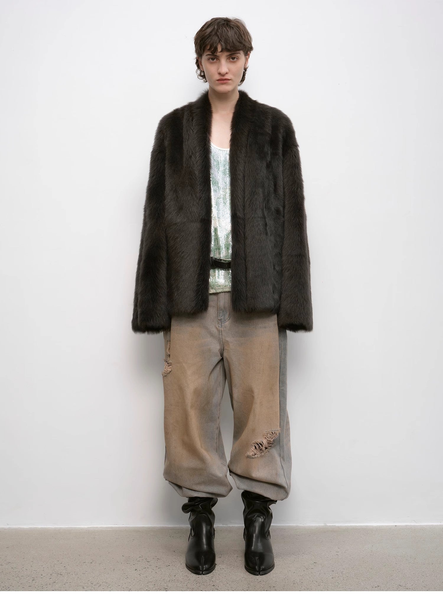 Person wearing a dark fur coat, light shirt, beige pants, and black boots against a white background