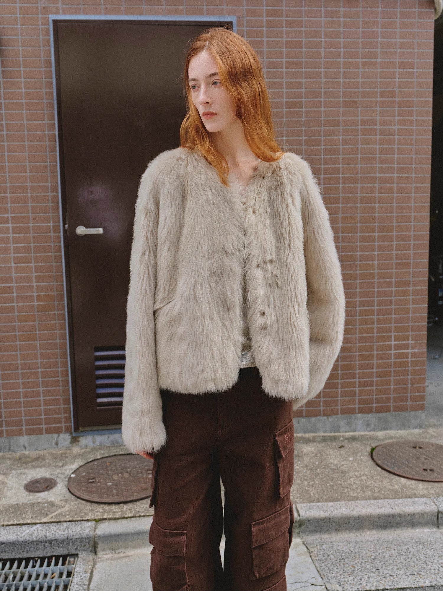 Person wearing a light gray fur jacket and brown pants standing in front of a tiled wall.