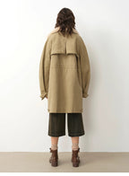 Person wearing a beige coat and dark pants against a white wall