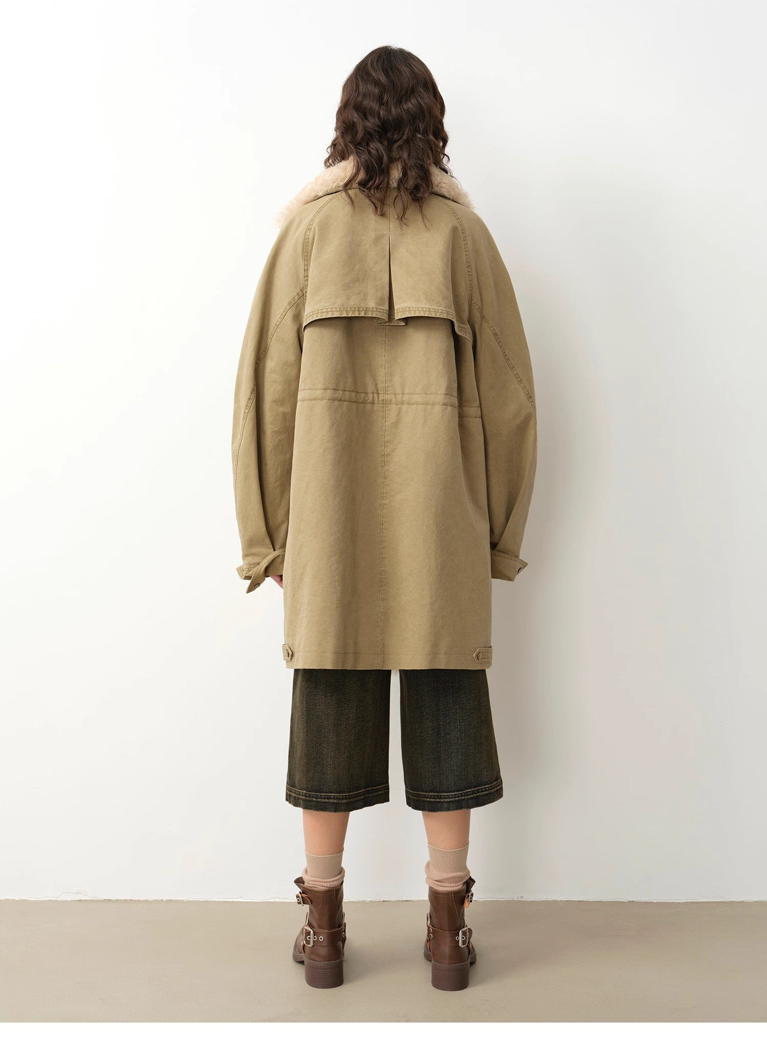 Person wearing a beige coat and dark pants against a white wall