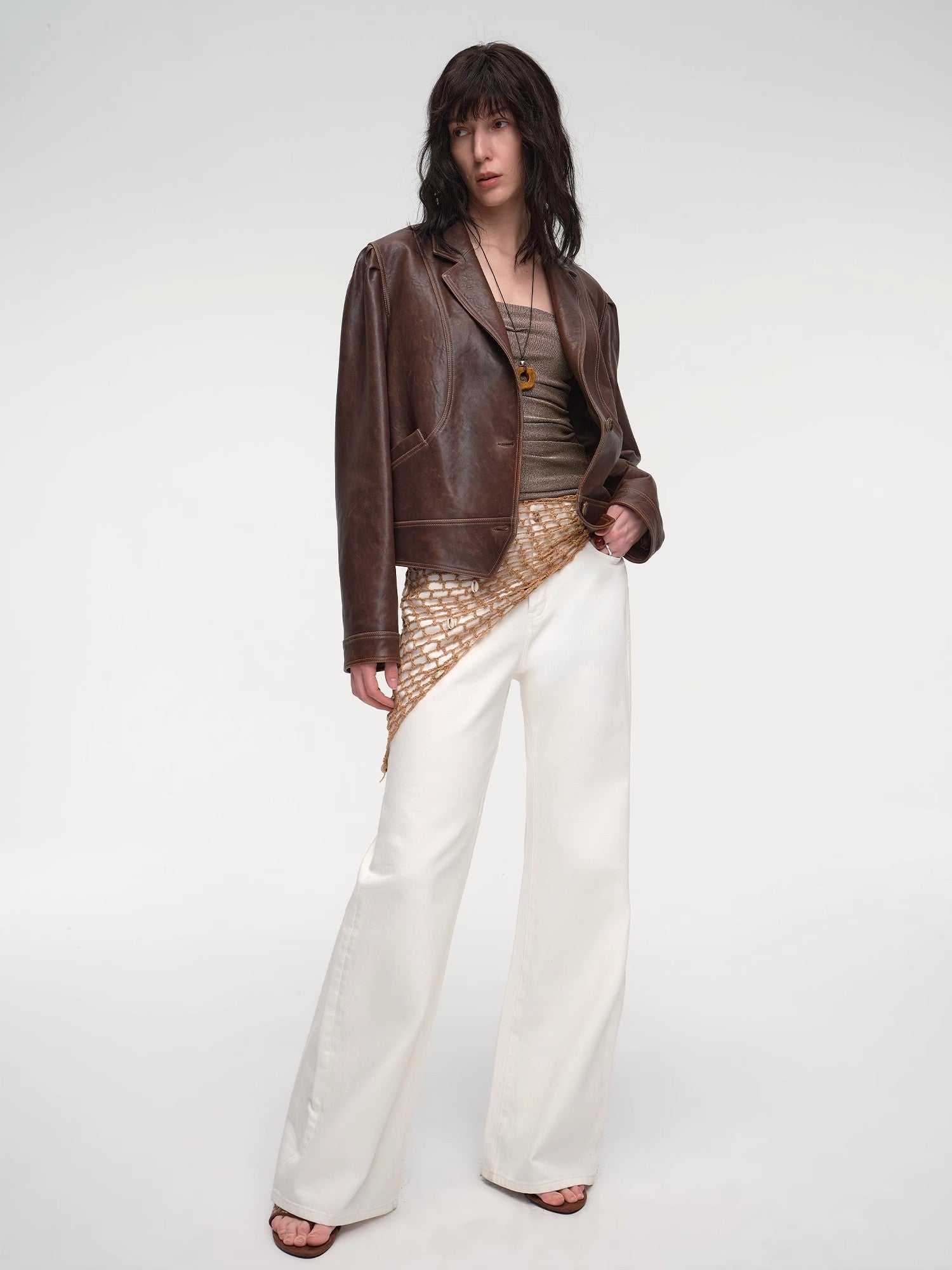 Woman wearing a brown leather jacket, patterned top, and white pants on a white background