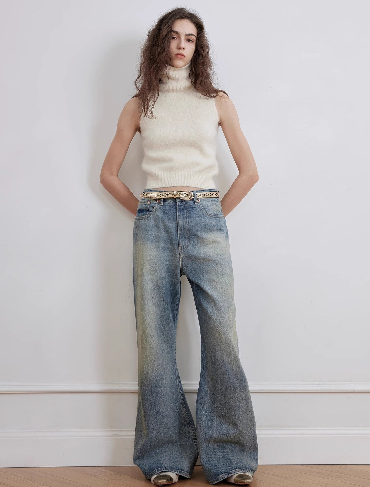 Woman wearing a cream sleeveless top and blue jeans against a white wall.