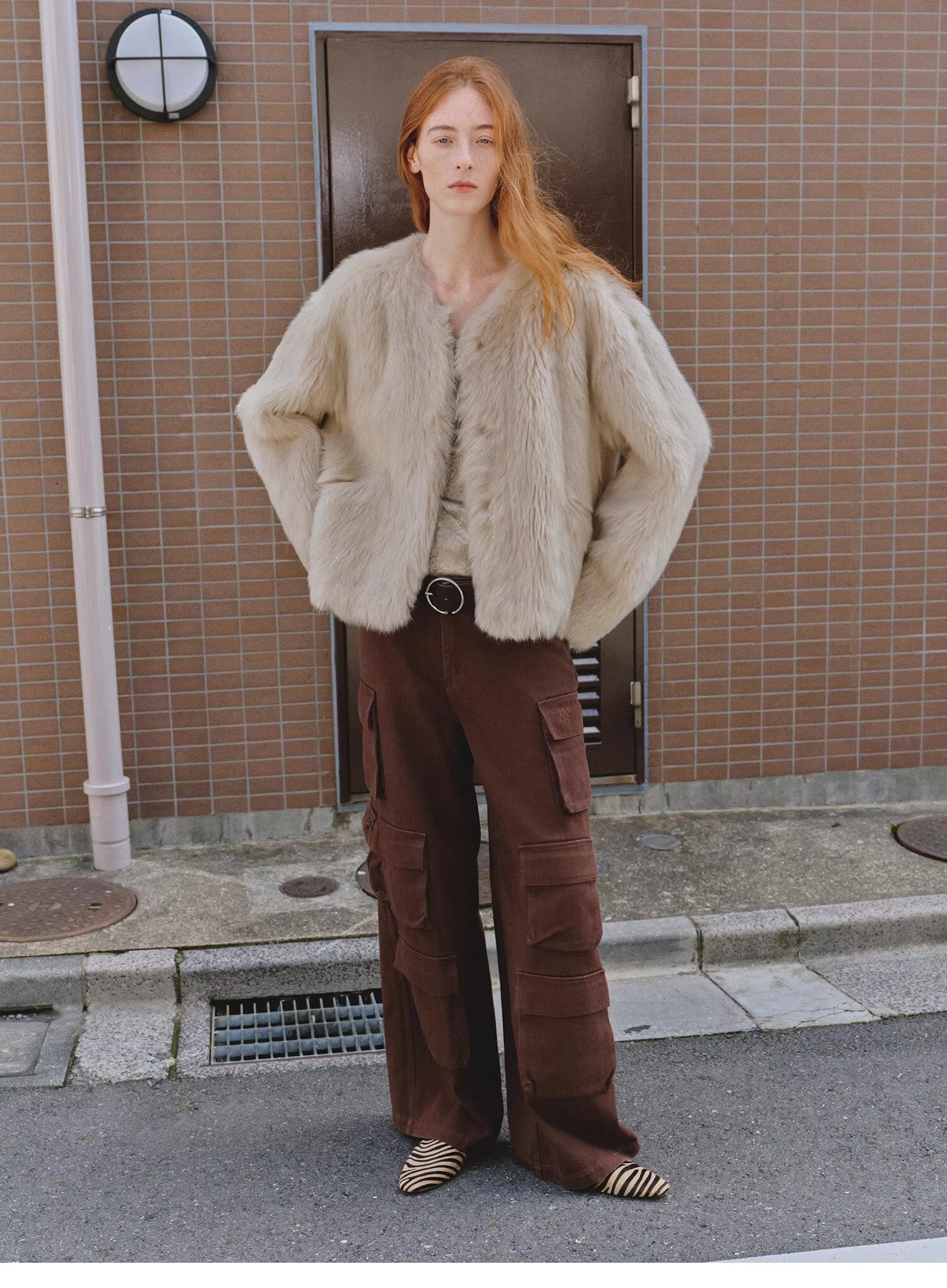 Person wearing a beige fur coat and brown pants standing on a street.