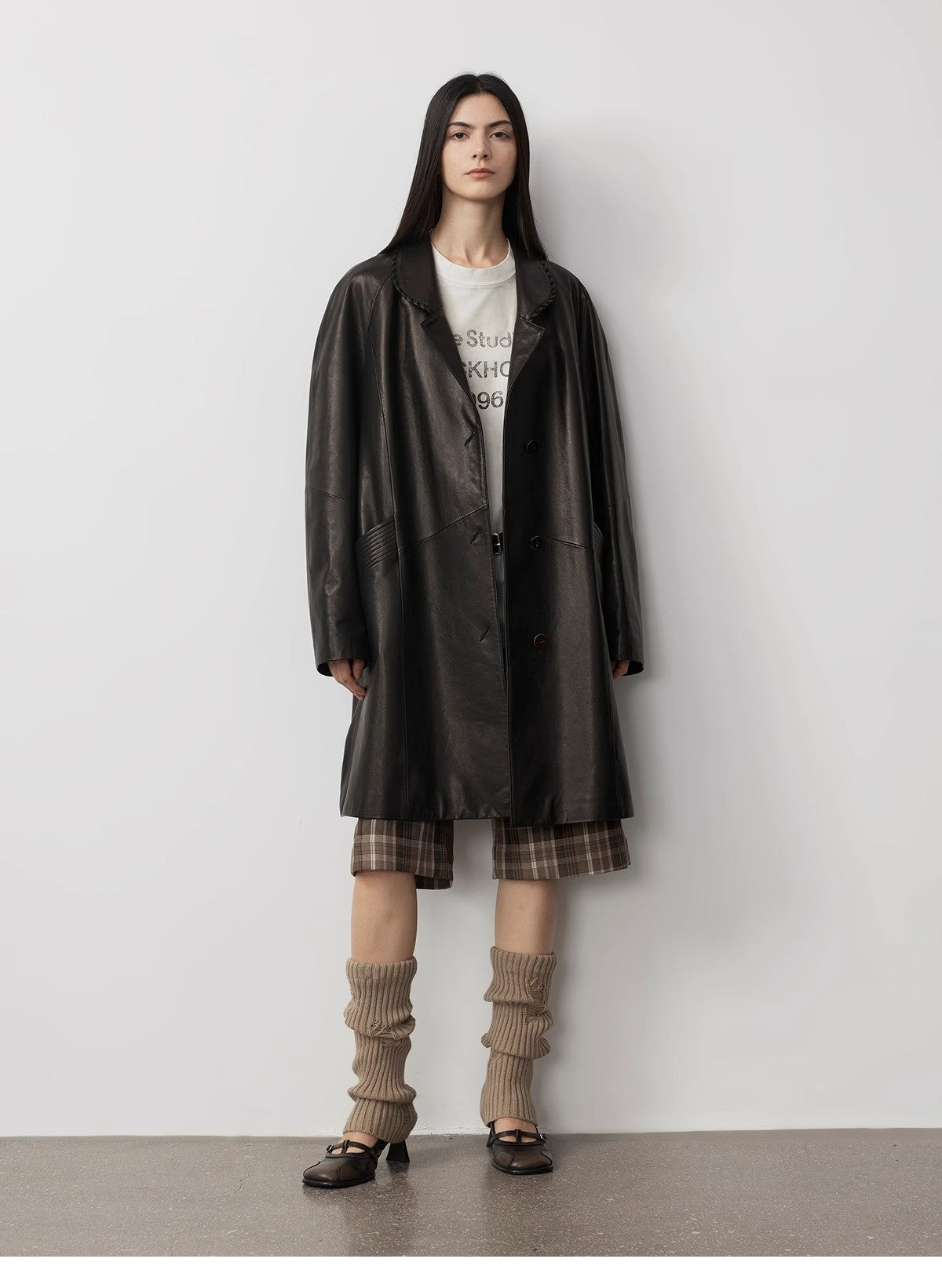 Person wearing a dark coat over a plaid dress with long socks and shoes on a plain background