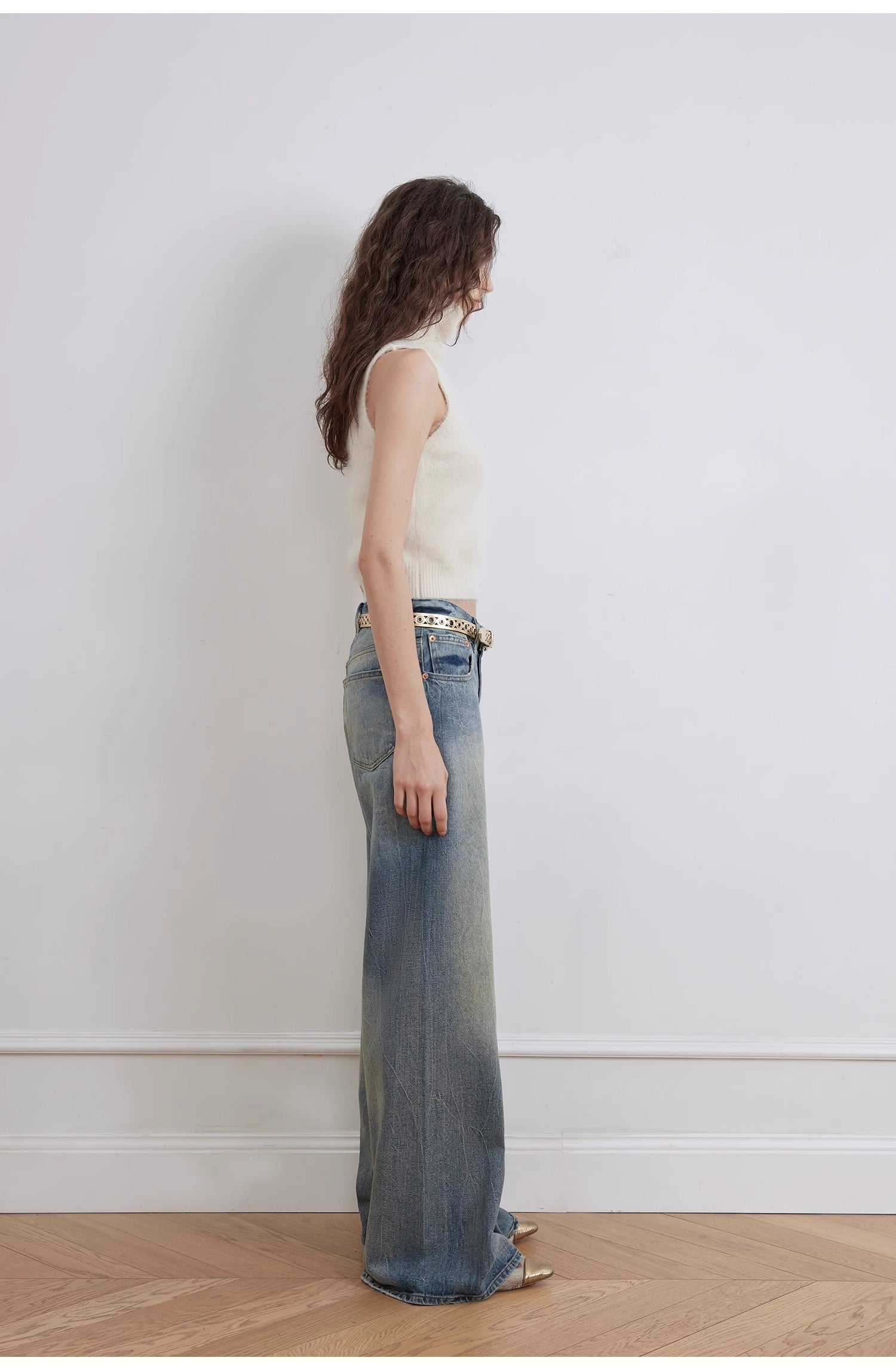 Woman wearing a white sleeveless top and blue jeans against a plain white wall.