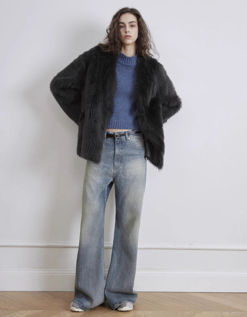 Person wearing a black fur coat, blue sweater, and jeans against a white wall.