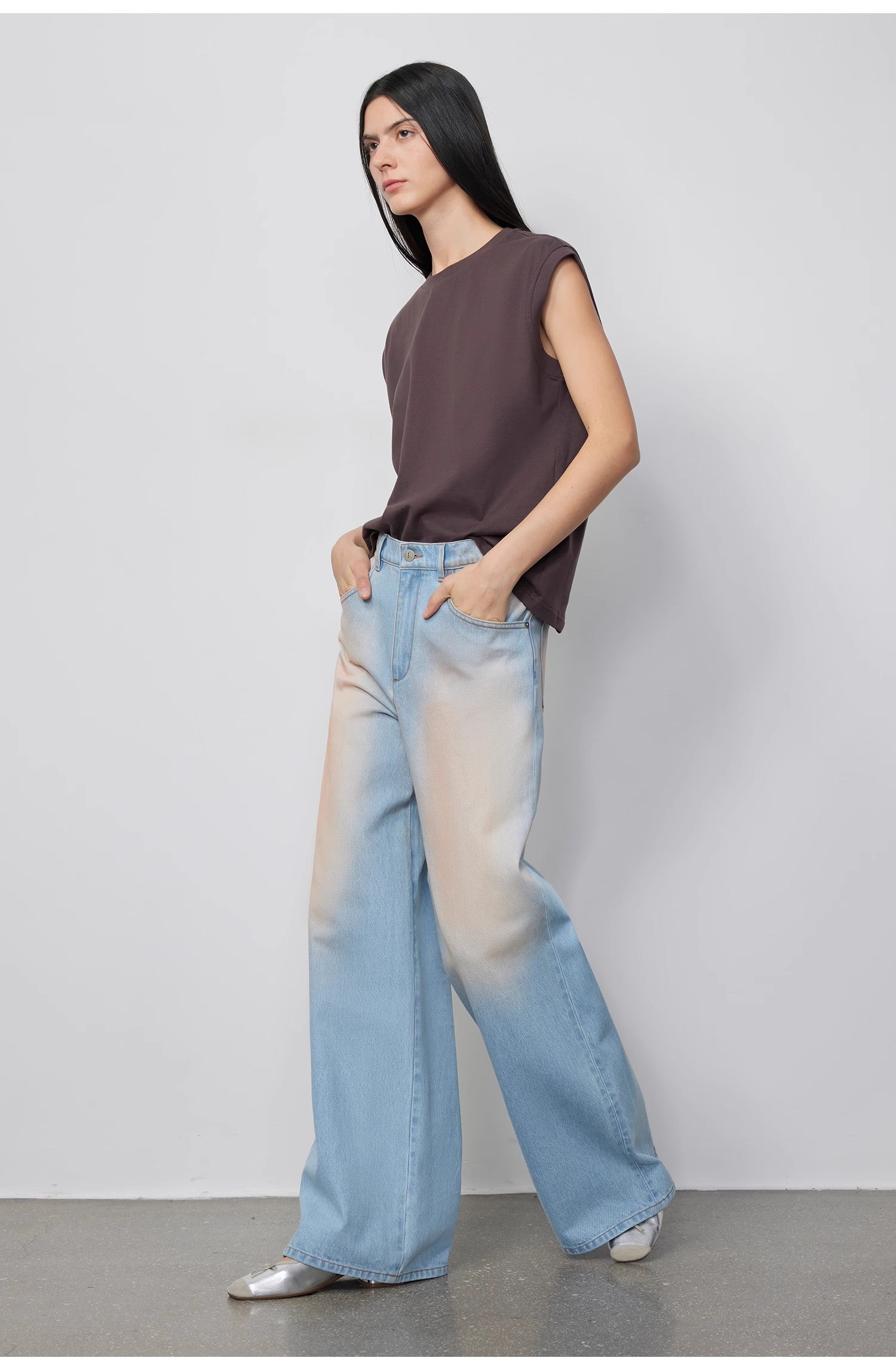 Woman wearing a sleeveless brown top and light blue jeans against a plain background