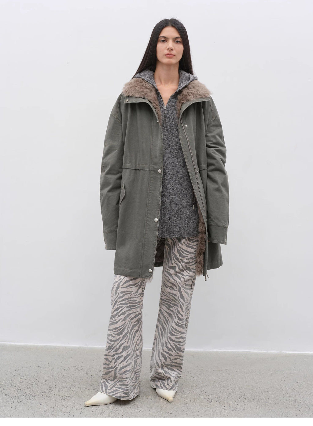 Person wearing a green coat with fur lining, gray sweater, and zebra print pants on a white background
