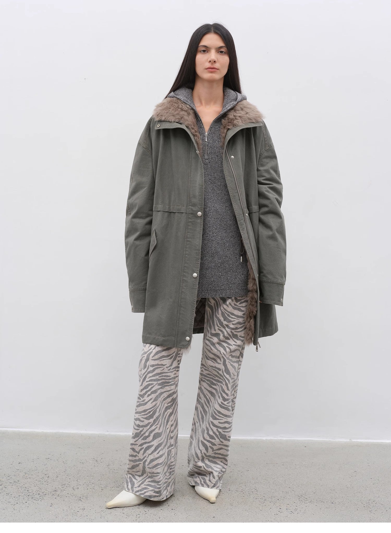 Person wearing a green coat with fur lining, gray sweater, and zebra print pants on a white background