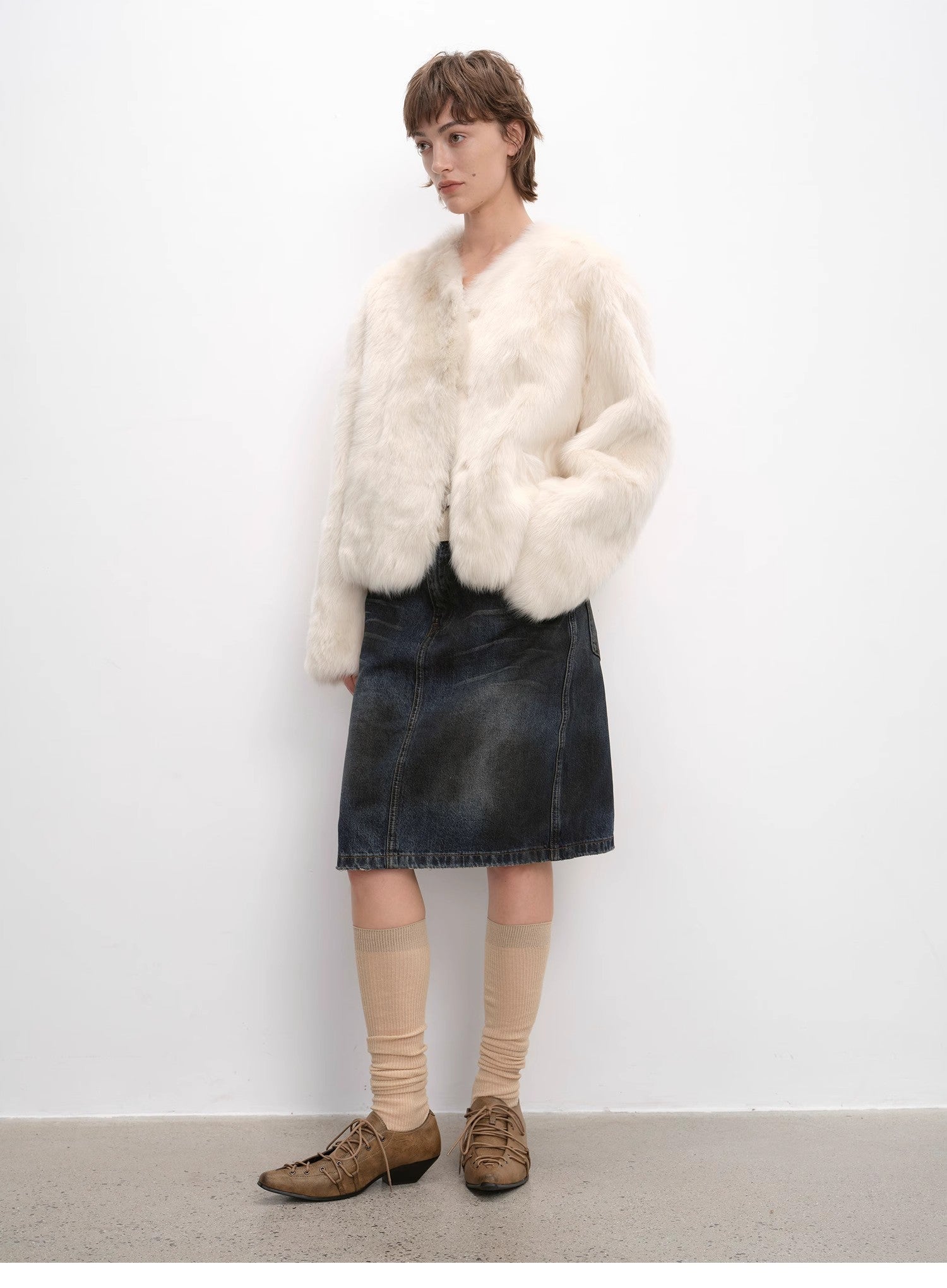 Person wearing a white fur coat, denim skirt, and brown shoes against a white background