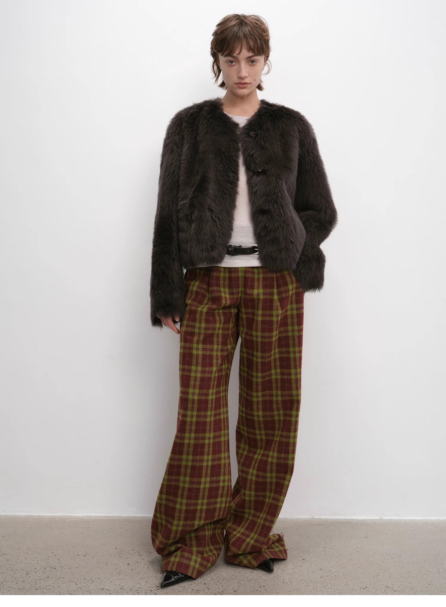 Person wearing a dark brown fur coat and plaid pants against a white wall.